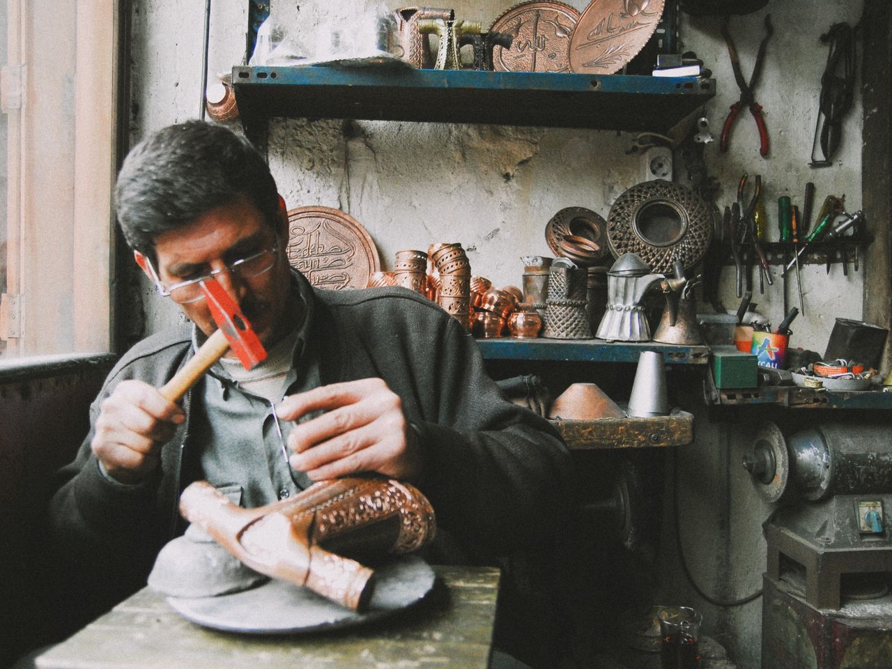 A skilled artisan sculpting copperware with a hammer in a traditional, tool-filled workshop.